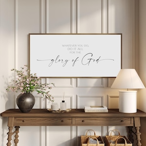 May include: A rectangular wall sign with a white background and a thin brown frame. The sign features black text that reads: WHATEVER YOU DO, DO IT ALL FOR THE glory of God. The text is centered with a mix of block lettering and cursive script.