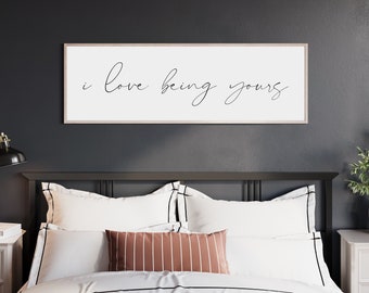 May include: A black framed wall sign with the text "i love being yours" in white cursive script, hanging above a bed with white bedding and a brown and white striped throw pillow. The bed is in a room with dark gray walls and a nightstand on each side.