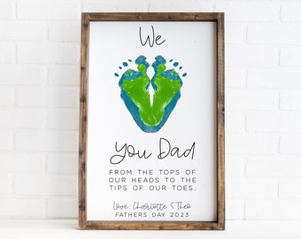May include: A framed white sign with a brown wooden frame. The sign features a heart-shaped design made of green and blue footprints. The text reads "We You Dad" and a message below. Signed "Love, Charlotte & Theo Fathers Day 2023".