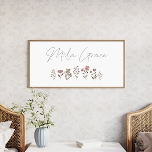 May include: A rectangular wall sign with a light wood frame featuring the text Mila Grace in black cursive script above a row of small, colourful botanical illustrations of various wildflowers in shades of pink, green, and brown.