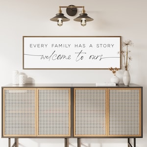 May include: A white sign with black text that reads "Every family has a story, welcome to ours". The sign is hanging on a white wall above a dark brown cabinet with a woven rattan front.