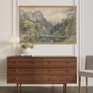 May include: A wooden dresser with six drawers and brass ring pulls. Above the dresser hangs a landscape painting of a mountain and river scene. A white lamp and vase of flowers sit on top of the dresser. A gray chair is to the right.