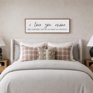 Puede incluir: Una escena de dormitorio con una cama con un edred&oacute;n gris claro y almohadas decorativas. Encima de la cama hay un letrero de madera con el texto "i love you more than yesterday, but not as much as tomorrow". Dos l&aacute;mparas negras est&aacute;n sobre mesitas de noche de madera.
