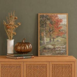 May include: A framed vertical print featuring a landscape painting of autumn trees with red and green foliage, including the text "fall is proof that change is beautiful". Beside it sits a white vase with dried grasses and a brown glass pumpkin.