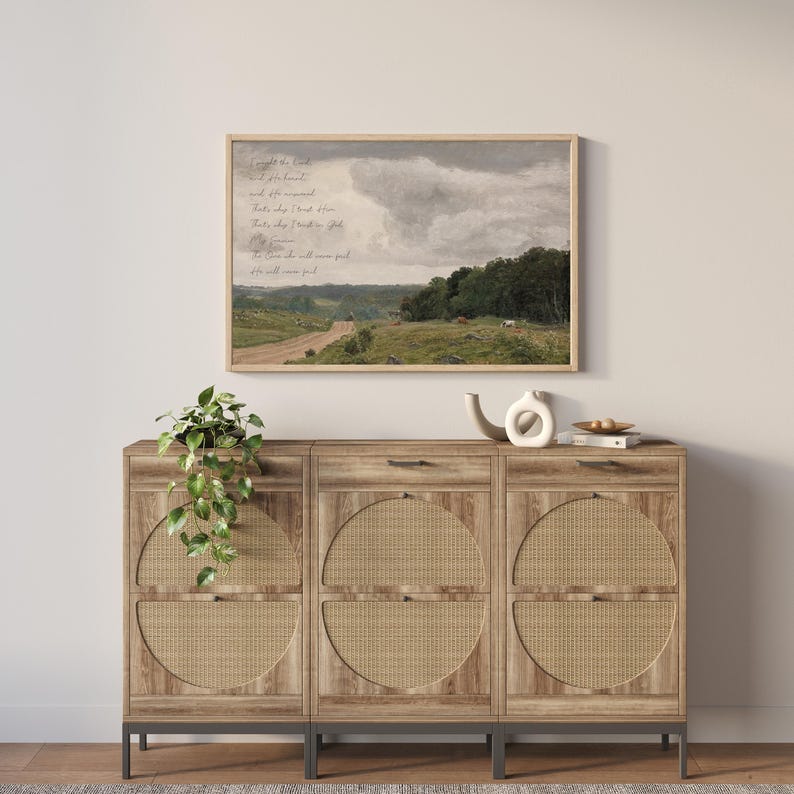 May include: A landscape wall art print in a light wood frame featuring a rolling green countryside, a dirt path, and a cloudy sky with cursive text overlay. Below sits a light brown wooden sideboard with three cabinets featuring woven rattan circular panels.
