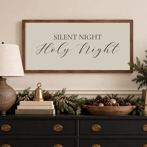 May include: A rectangular wall sign with a dark wood frame and a light beige background featuring the black text SILENT NIGHT Holy Night in a combination of serif and script fonts, centered horizontally and mounted above a dark console table.