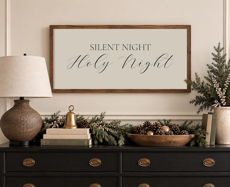 May include: A rectangular wall sign with a dark wood frame and a light beige background featuring the black text SILENT NIGHT Holy Night in a combination of serif and script fonts, centered horizontally and mounted above a dark console table.
