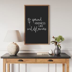 以下が含まれることがあります： 「Spread Kindness Like Wildflowers」と書かれた白い文字が入った、額入りの黒い看板。看板は茶色の木製フレームに入っており、木製のテーブルに飾られています。ランプ、花瓶、本もテーブルにあります。