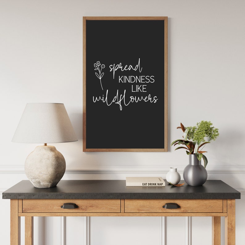 以下が含まれることがあります： 「Spread Kindness Like Wildflowers」と書かれた白い文字が入った、額入りの黒い看板。看板は茶色の木製フレームに入っており、木製のテーブルに飾られています。ランプ、花瓶、本もテーブルにあります。