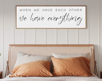 Puede incluir: Un letrero de madera con el texto "When we have each other we have everything" en letra cursiva negra. El letrero está colgado sobre una cama con una cabecera beige y almohadas naranjas y a rayas.