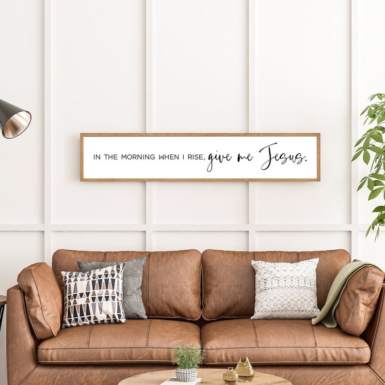 May include: A wooden framed sign with the text "In the morning when I rise, give me Jesus." The sign is hanging on a white wall above a brown leather couch.