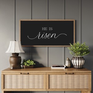 May include: A black framed sign with white text that reads "He is risen" hanging on a gray wall above a wooden console table with two drawers. The table has a woven rattan front and is decorated with a lamp, a potted plant, and a stack of books.