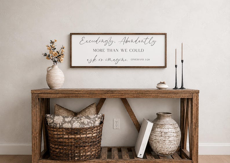 Op de afbeelding: Een rustieke houten console met decoratieve items. Boven de tafel hangt een ingelijst bord met scripttekst. De tafel bevat een vaas met bloemen, een gevlochten mand, een keramische vaas, kaarsen en een klein schaaltje.