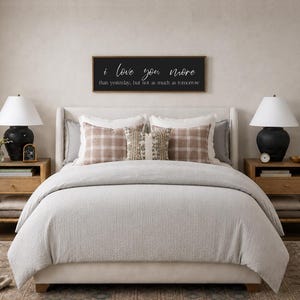 Puede incluir: Una escena de dormitorio con una cama con un edred&oacute;n gris claro y almohadas decorativas. Un letrero enmarcado en negro sobre la cama dice "I love you more than yesterday, but not as much as tomorrow." Dos l&aacute;mparas negras se encuentran en mesitas de noche de madera.
