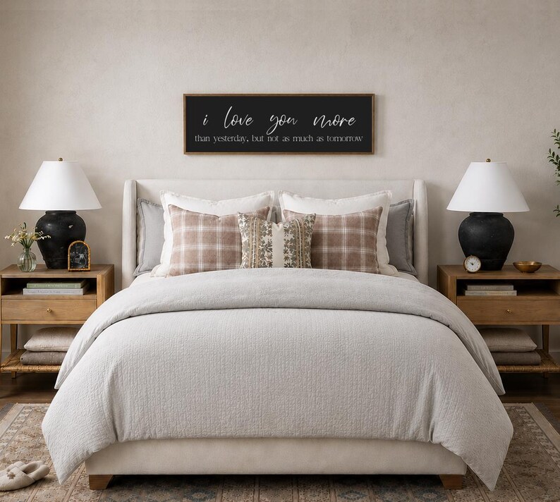 Puede incluir: Una escena de dormitorio con una cama con un edred&oacute;n gris claro y almohadas decorativas. Un letrero enmarcado en negro sobre la cama dice "I love you more than yesterday, but not as much as tomorrow." Dos l&aacute;mparas negras se encuentran en mesitas de noche de madera.