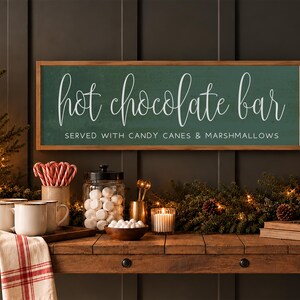 May include: A rectangular wooden-framed sign with a dark green background and white text that reads hot chocolate bar SERVED WITH CANDY CANES & MARSHMALLOWS in a mix of cursive and block lettering, mounted on a dark paneled wall.