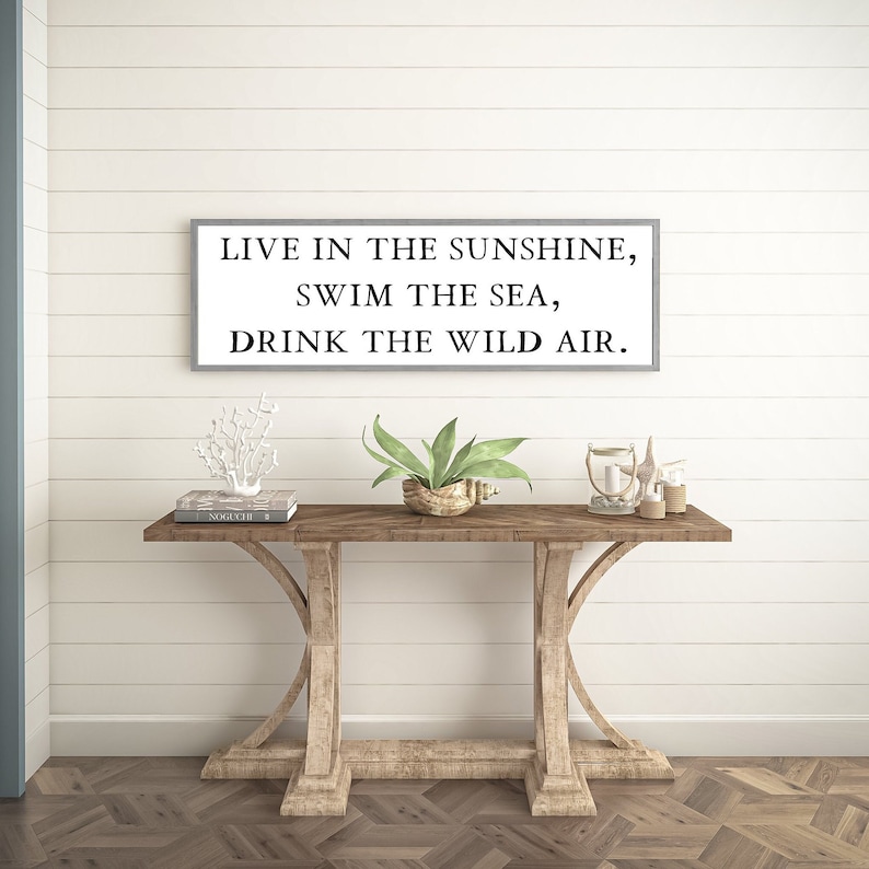 May include: A white framed sign with the text "Live in the sunshine, swim the sea, drink the wild air." The sign is hanging on a white shiplap wall above a wooden console table.