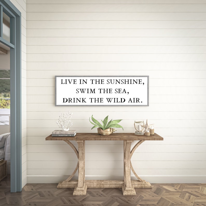 May include: A wooden console table with a white sign above it that reads "Live in the sunshine, swim the sea, drink the wild air."