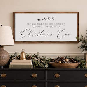 May include: A rectangular wooden framed sign with a white background featuring a black silhouette of Santa in a sleigh pulled by reindeer and the text: MAY YOU NEVER BE TOO GROWN UP TO SEARCH THE SKIES ON Christmas Eve.