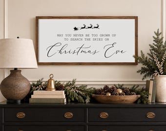 May include: A rectangular wall sign with a dark wood frame and a white background. The sign features a black silhouette of Santa in a sleigh with reindeer, and the text: MAY YOU NEVER BE TOO GROWN UP TO SEARCH THE SKIES ON Christmas Eve.