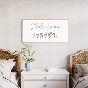 May include: A rectangular wall sign with a light wood frame featuring the text Mila Grace in black cursive script above a row of small, muted-toned botanical illustrations including various flowers and leaves in shades of pink, green, and brown.