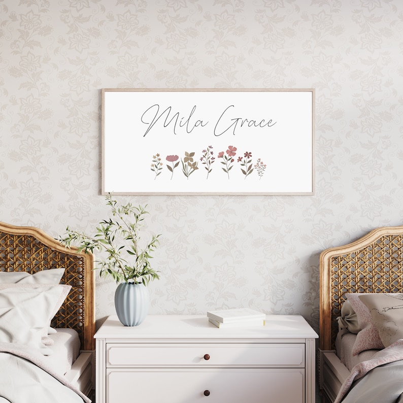 May include: A rectangular wall sign with a light wood frame featuring the text Mila Grace in black cursive script above a row of small, muted-toned botanical illustrations including various flowers and leaves in shades of pink, green, and brown.