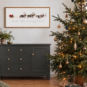 Puede incluir: Un letrero de pared rectangular con marco de madera que presenta un gr&aacute;fico de Santa Claus en un trineo tirado por renos y el texto Santa Claus is coming to town en letras cursivas negras sobre un fondo blanco.