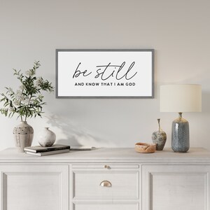 May include: A rectangular wall sign with a gray frame and a white background. The text "be still" is written in a black cursive script, with the words "AND KNOW THAT I AM GOD" printed in a smaller, black, sans-serif, all-caps font below it.