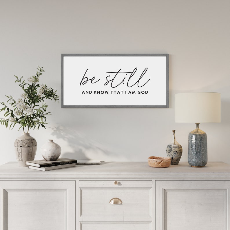 May include: A rectangular wall sign with a gray frame and a white background. The text "be still" is written in a black cursive script, with the words "AND KNOW THAT I AM GOD" printed in a smaller, black, sans-serif, all-caps font below it.