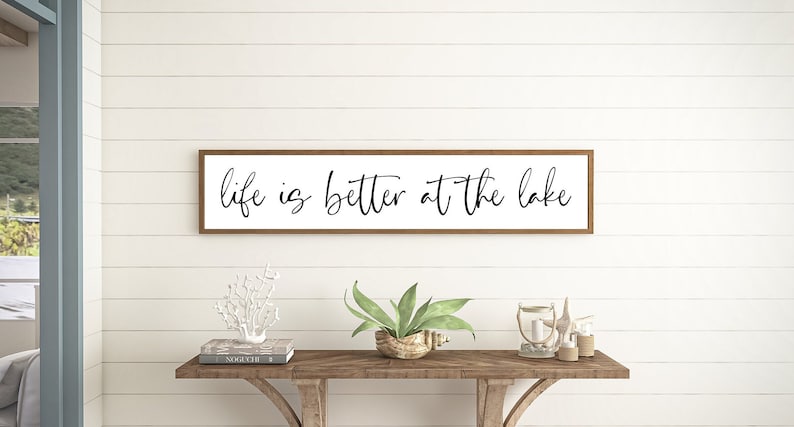 May include: A rectangular wall sign with a thin brown wooden frame and a white background. The text "life is better at the lake" is printed in a black, cursive script font across the center of the sign, spanning the length of the frame.