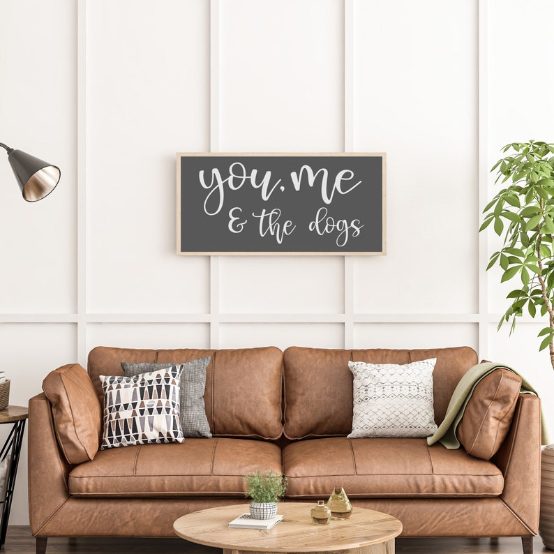 May include: A rectangular wall sign with a light wood frame and a dark gray background featuring the white cursive text "you, me & the dogs" centered on the surface. The sign is mounted on a white paneled wall above a brown leather sofa.