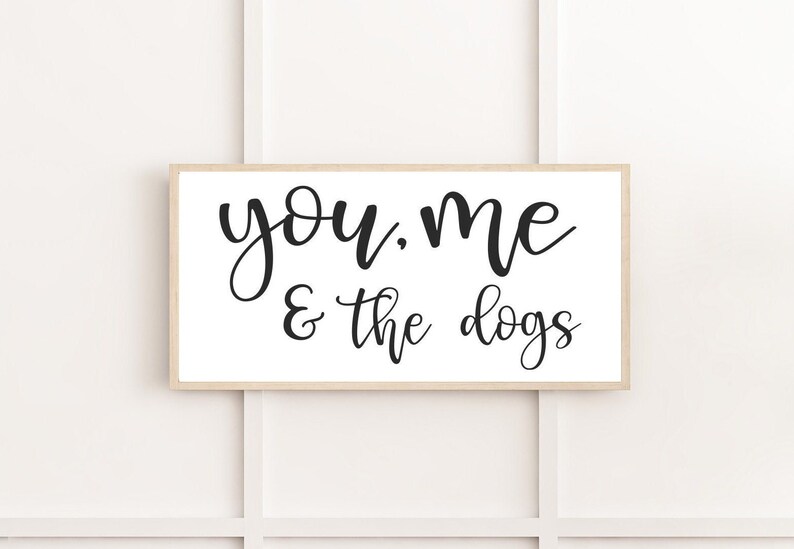May include: A rectangular wall sign with a light wood frame and a white background. The text "you, me & the dogs" is printed in black, cursive lettering across the center of the sign. The design is simple, featuring clean lines and high contrast.