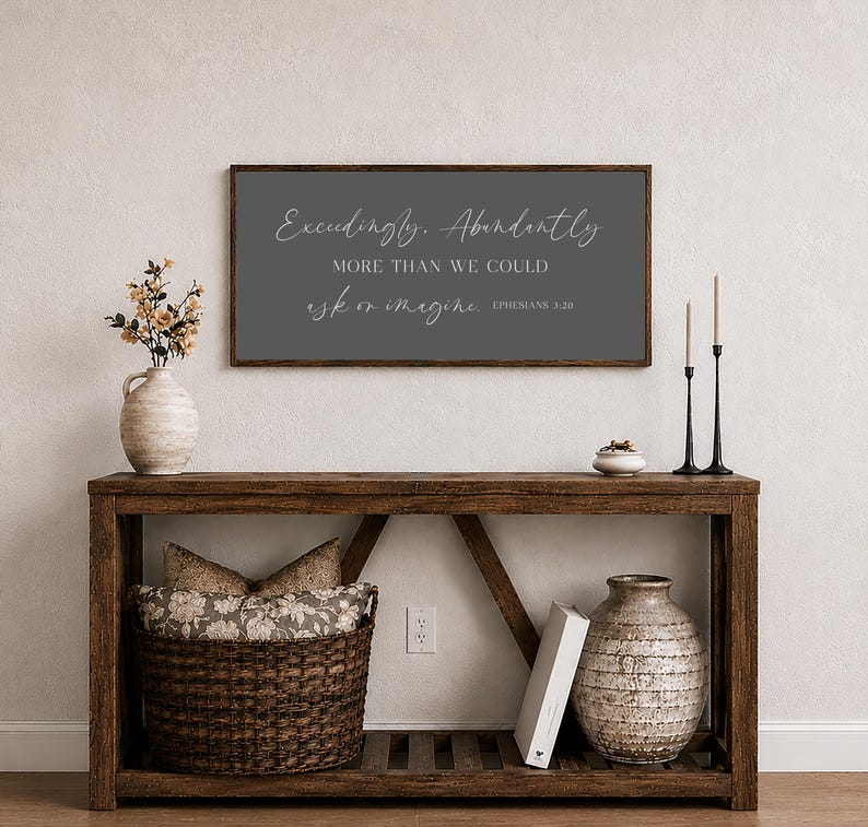 Op de afbeelding: Een houten console met een donkergrijs omlijst bord met de tekst "Exceedingly, Abundantly, More Than We Could Ask or Imagine, Ephesians 3:20." De tafel heeft een gevlochten mand, een vaas en decoratieve items.