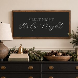 May include: A rectangular wall sign with a dark brown wooden frame and a black background featuring the white text "SILENT NIGHT" in a sans-serif font above the words "Holy Night" written in a white, elegant, flowing cursive script.
