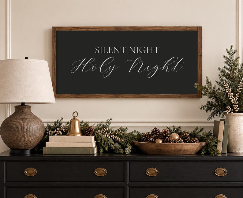 May include: A rectangular wall sign with a dark brown wooden frame and a black background featuring the white text "SILENT NIGHT" in a sans-serif font above the words "Holy Night" written in a white, elegant, flowing cursive script.