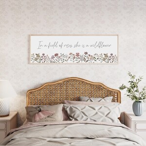 Puede incluir: Un letrero de pared rectangular con marco de madera clara que presenta el texto cursivo negro In a field of roses, she is a wildflower sobre una fila horizontal de flores silvestres ilustradas en tonos apagados de rosa, verde y marr&oacute;n.