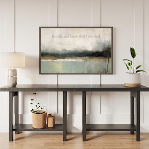 May include: A framed landscape art print with the text "Be still, and know that I am God." The artwork features a muted color palette of blues, greens, and browns. The print is displayed above a dark gray console table with a lamp and potted plants.