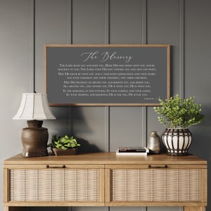 Peut inclure: Une &oelig;uvre d'art encadr&eacute;e avec le texte "The Blessing" en &eacute;criture &eacute;l&eacute;gante. L'&oelig;uvre est expos&eacute;e au-dessus d'une console en bois avec une lampe, de la verdure et des objets d&eacute;coratifs. Le mur est peint en gris neutre.