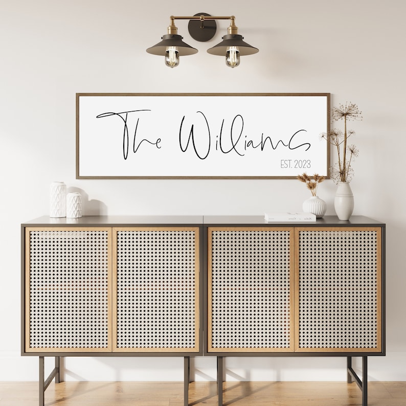 May include: A rectangular wall sign with a dark wood frame and a white background featuring the black text The Williams and EST. 2023 in a script and sans-serif font. The sign is mounted on a wall above a dark cabinet with four cane doors.