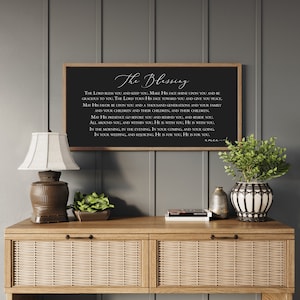 Peut inclure: Un panneau noir encadr&eacute; avec le texte "The Blessing" en &eacute;criture blanche. Le panneau est expos&eacute; sur une console en bois avec une lampe, des plantes et des objets d&eacute;coratifs. Le mur derri&egrave;re la table est peint en gris.
