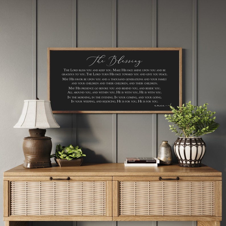 Peut inclure: Un panneau noir encadr&eacute; avec le texte "The Blessing" en &eacute;criture blanche. Le panneau est expos&eacute; sur une console en bois avec une lampe, des plantes et des objets d&eacute;coratifs. Le mur derri&egrave;re la table est peint en gris.