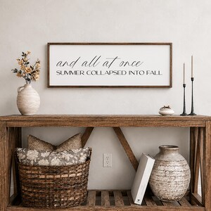 May include: A rustic wooden console table with decorative items. A framed sign with the text "and all at once SUMMER COLLAPSED INTO FALL" hangs above. The table holds a vase with flowers, a woven basket, and a large vase. Two black candlesticks with candles are on the right.