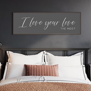 May include: A bedroom with a black headboard and a white bedspread. A framed sign with the text "I love your love the most" hangs above the bed.