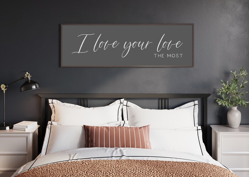 May include: A bedroom with a black headboard and a white bedspread. A framed sign with the text "I love your love the most" hangs above the bed.