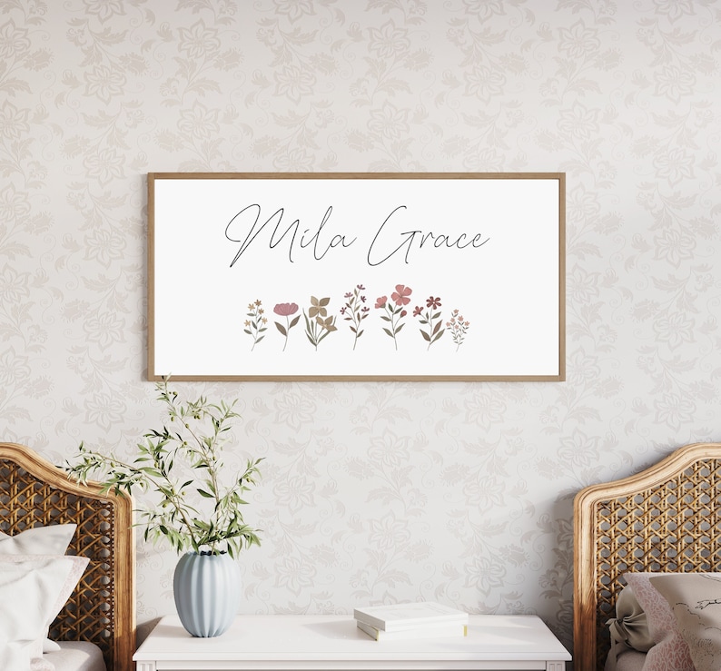 May include: A rectangular wall sign with a light wood frame featuring the text Mila Grace in black cursive script above a row of small, colourful botanical illustrations of various wildflowers in shades of pink, green, and brown.