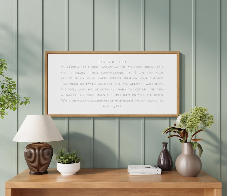 May include: A framed rectangular sign with the text "Love the Lord" in a wooden frame. The sign is displayed on a wooden table with a lamp, a small plant, and decorative vases.