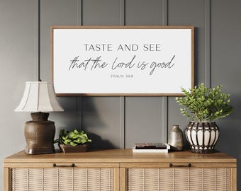 May include: A rectangular wall sign with a light wood frame featuring the black text TASTE AND SEE that the Lord is good PSALM 34:8 on a white background. The sign is displayed above a wooden console table with woven cabinet doors.