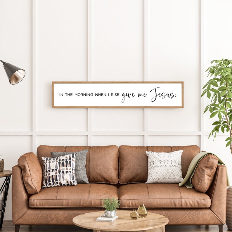 May include: A wooden sign with the text "In the morning when I rise, give me Jesus." The sign is hanging on a white wall above a brown leather couch.