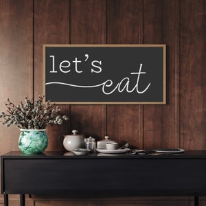 May include: A black framed sign with the words "let's eat" in white script hangs above a black cabinet. A green and blue vase with a floral arrangement sits to the left of the sign. A teapot, dishes, and other items are on the cabinet.