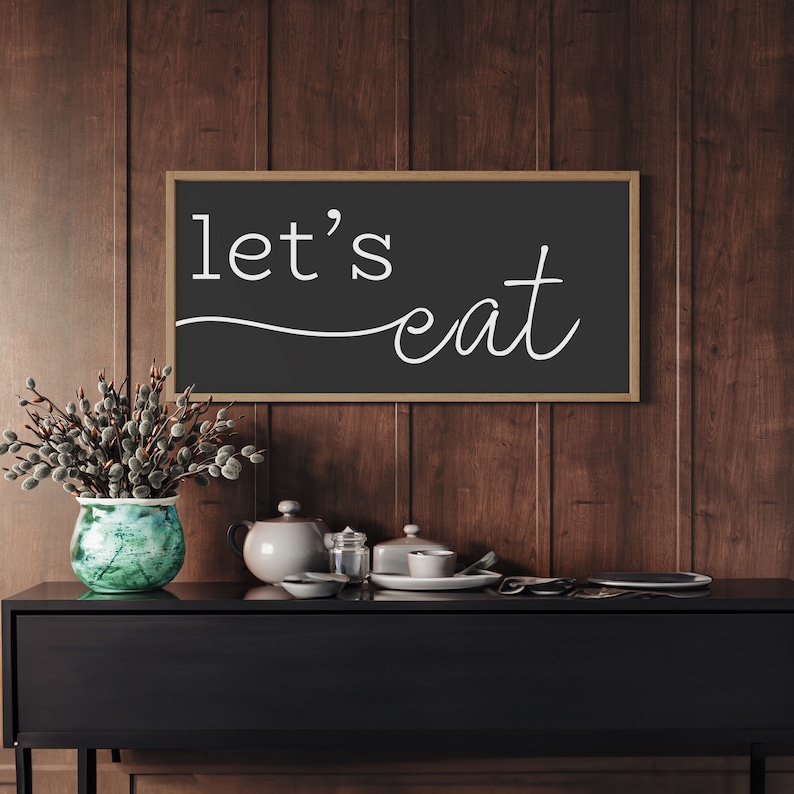 May include: A black framed sign with the words "let's eat" in white script hangs above a black cabinet. A green and blue vase with a floral arrangement sits to the left of the sign. A teapot, dishes, and other items are on the cabinet.